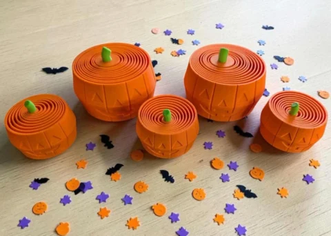 May include: Five orange pumpkin-shaped containers with green stems and carved jack-o'-lantern faces. The tops feature a concentric circular design. The set is surrounded by small, scattered confetti pieces in the shapes of bats, stars, and pumpkins.