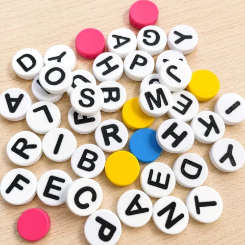 May include: A collection of round, white plastic beads featuring black printed letters of the alphabet, interspersed with solid, unprinted circular beads in vibrant shades of pink, yellow, and blue, scattered across a light wood surface.