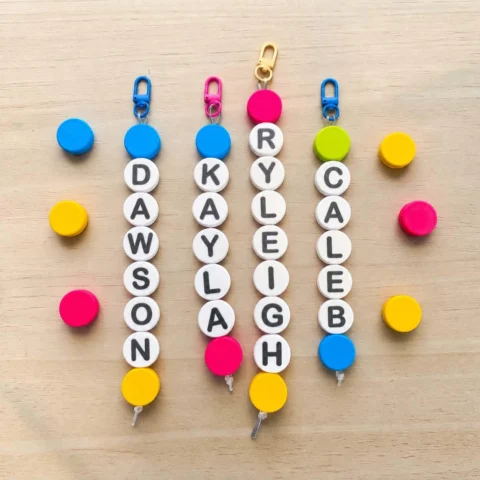May include: Four vertical keychains featuring white circular beads with black letters spelling the names Dawson, Kayla, Ryleigh, and Caleb. Each keychain has a colorful silicone bead at the top and bottom, with a metal clasp attached to the top.