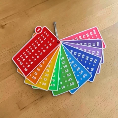 May include: A set of multi-colored multiplication table flashcards arranged in a fan shape and held together by a silver metal ring. Each rectangular card features white text displaying multiplication equations from 1 to 10, organized by color.