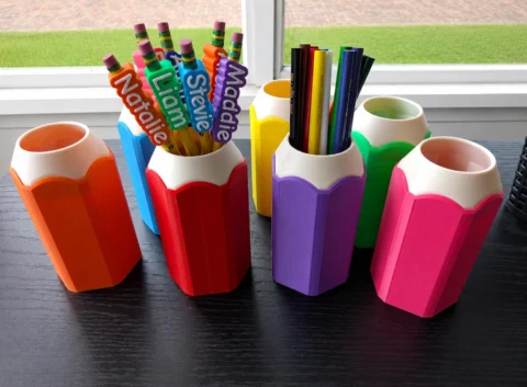 May include: A collection of hexagonal pencil-shaped pen holders in orange, red, purple, pink, yellow, blue, and green. Several pencils with personalized name toppers reading Natalie, Liam, Stevie, and Maddie are placed inside the containers.