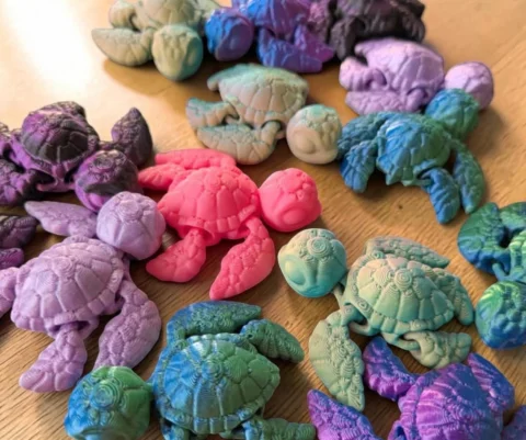 May include: A collection of articulated sea turtle figurines in various colors including pink, purple, blue, green, and gradient shades. Each turtle features a textured shell, movable flippers, and a detailed head, scattered across the frame.