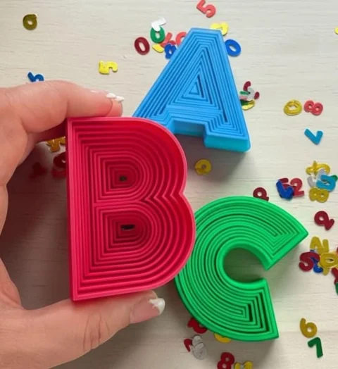 May include: Three plastic cookie cutters shaped like the letters A, B, and C. The letter A is light blue, the letter B is bright pink, and the letter C is green. Each letter features a series of concentric, raised ridges on its top surface.