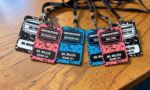 May include: A collection of rectangular lanyards featuring composition notebook patterns in black, pink, and blue. Each tag displays the text COMPOSITION NOTEBOOK, HALL PASS or RESTROOM PASS, and MS. WILCOX ROOM 101, attached to black fabric neck straps.