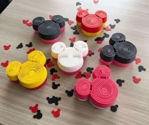 May include: Six three-dimensional mouse-shaped containers in red, yellow, black, white, and pink, each featuring a concentric circular pattern on the top surface. Small mouse-shaped confetti pieces in red and black are scattered around the containers.
