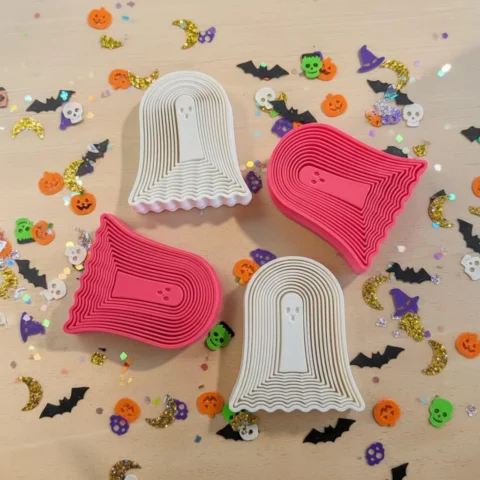 May include: Four ghost-shaped cookie cutters arranged in a square pattern. Two cutters are white and two are bright pink. Each cutter features a central ghost figure surrounded by concentric, wavy, tiered layers that create a dimensional effect.