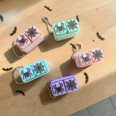 May include: Five rectangular pill cases in pastel pink, mint green, light blue, soft pink, and lavender. Each case features two compartments topped with black raised spider and spiderweb designs and includes a silver-toned metal ball chain.