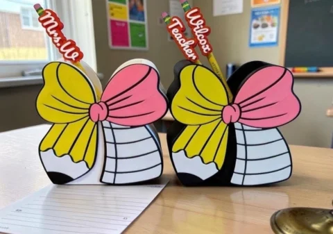May include: Two pencil holders shaped like bows with yellow pencil patterns and pink centers. One holder is white, the other is black. Each contains a pencil with a custom topper reading Mrs. U or Wilcox Teacher in red script lettering.
