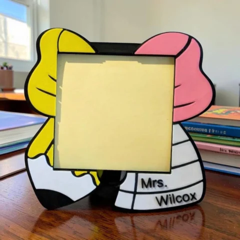 May include: A square photo frame featuring a stylized pencil design with a yellow top, a pink eraser section, and a white body. The frame includes black outlines and the text Mrs. Wilcox printed in black on the lower right side of the white pencil base.