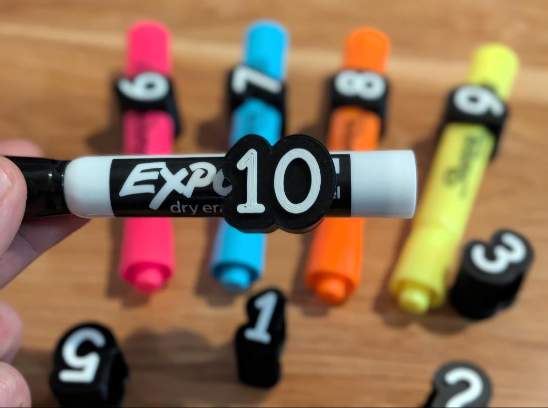 May include: A white Expo dry erase marker featuring a black clip with the number 10 in white. In the background, there are pink, blue, orange, and yellow markers with black clips numbered 6, 7, 8, and 6, along with loose clips numbered 5, 1, 3, and 2.