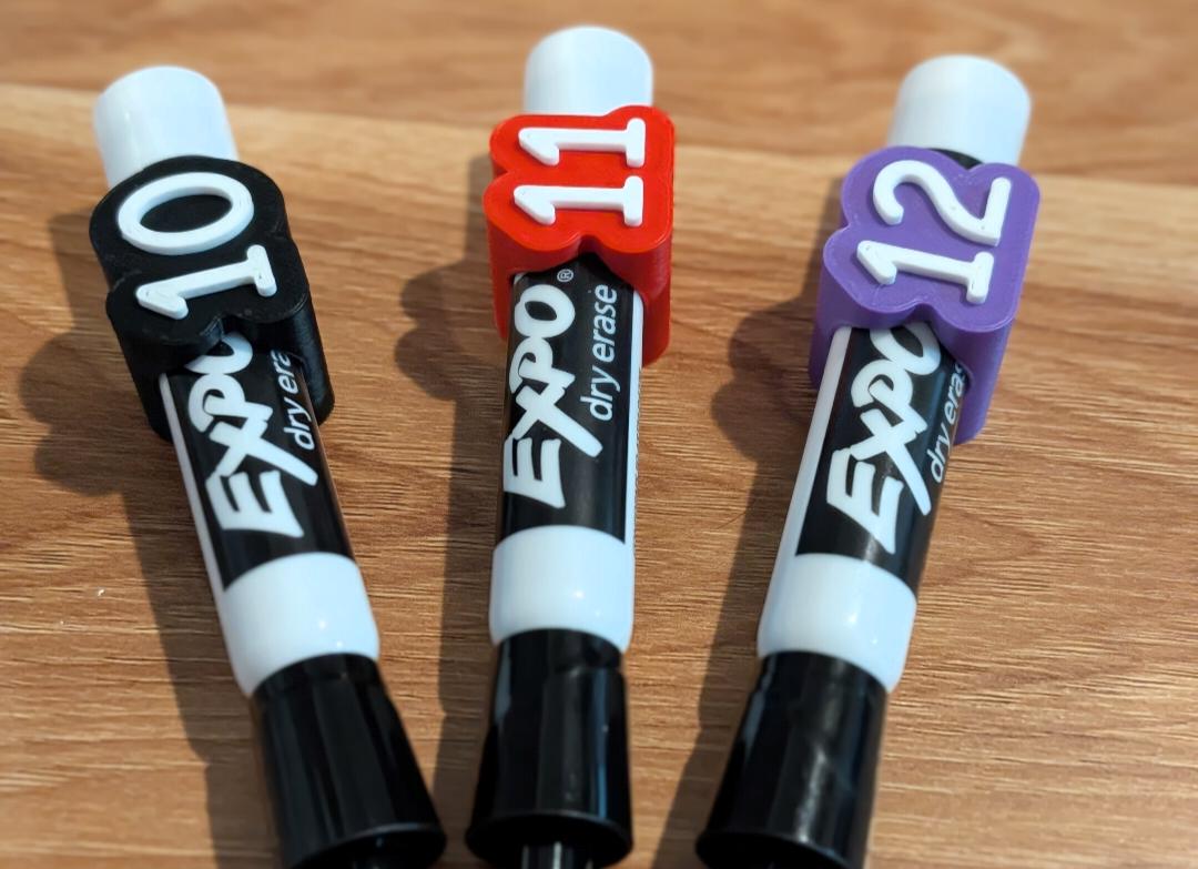 May include: Three Expo dry erase markers with custom plastic clips attached to the caps. The clips are black, red, and purple, featuring the white raised numbers 10, 11, and 12 respectively. Each marker has a white barrel with black text.