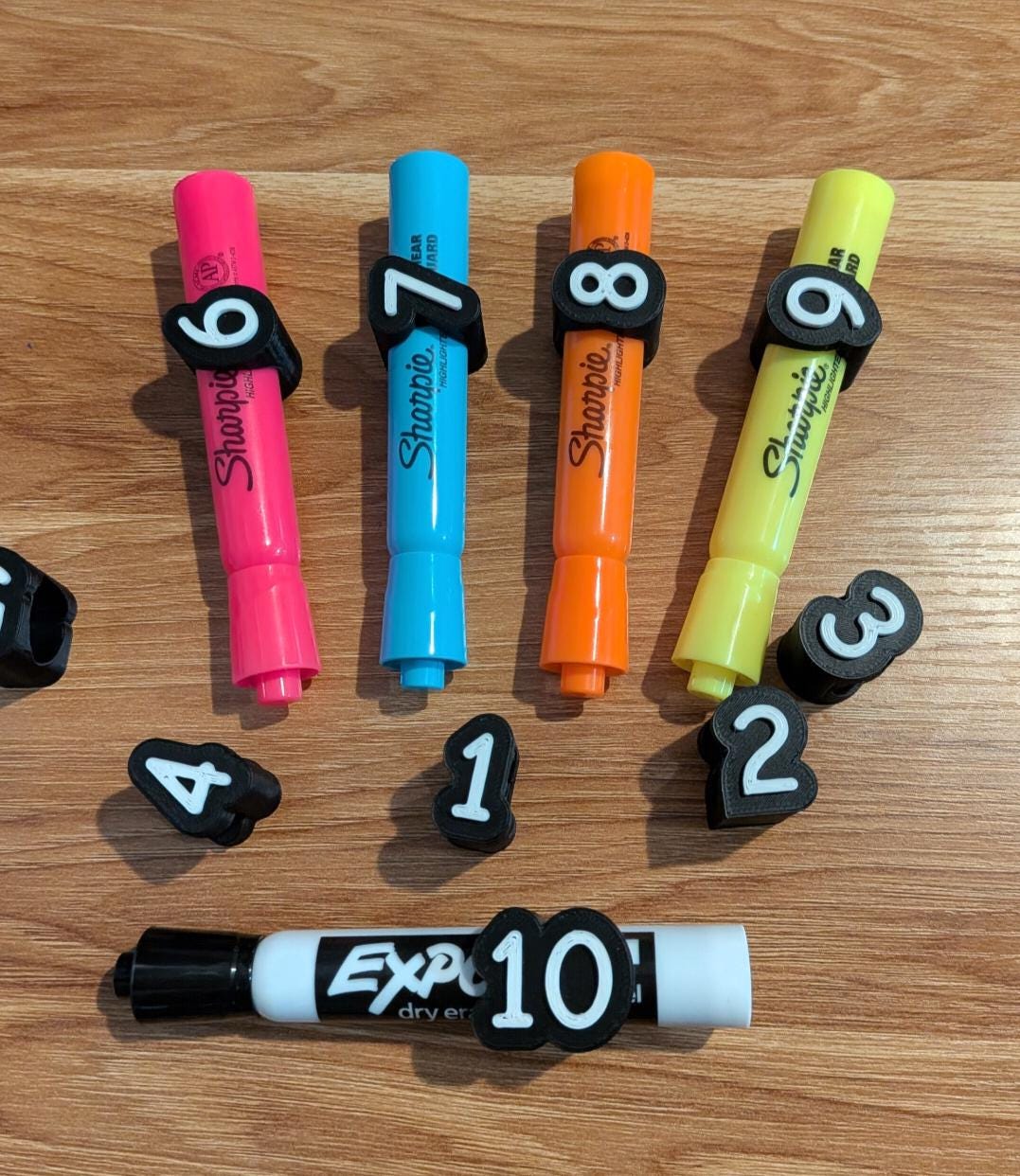 May include: A collection of four colorful Sharpie highlighters in pink, blue, orange, and yellow, each fitted with a black clip labeled with numbers 6, 7, 8, and 9. Below, a white Expo dry erase marker features a black clip labeled 10.
