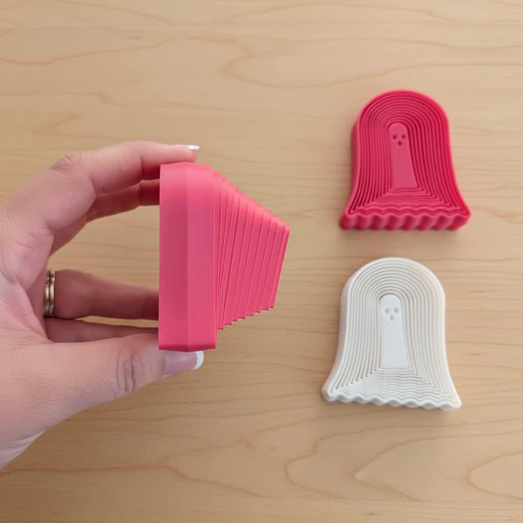 May include: Three ghost-shaped cookie cutters, one held in profile showing a thick pink side, and two flat on the surface in pink and white. Each cutter features a central ghost figure with three small circular eyes and mouth cutouts.