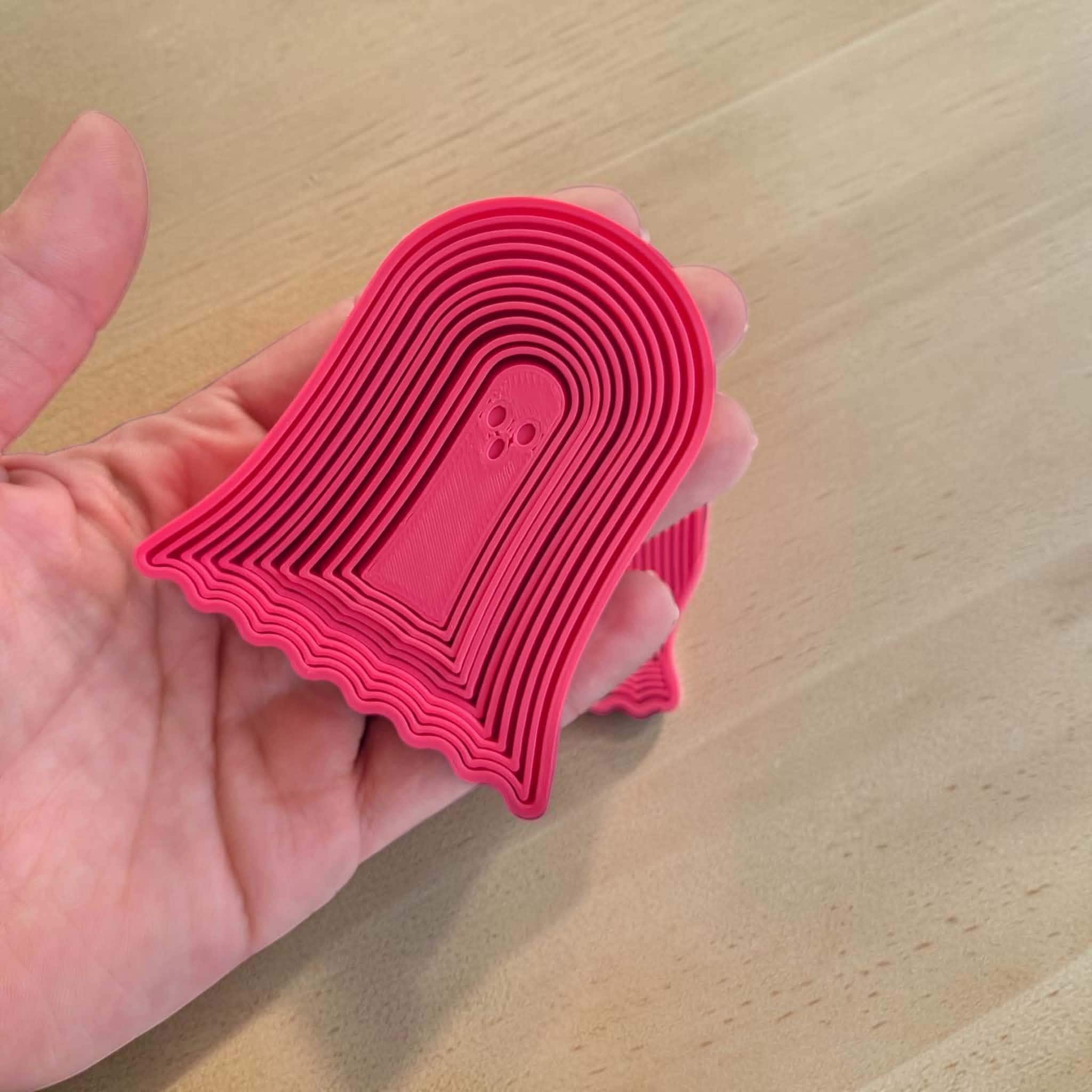 May include: A bright pink, ghost-shaped cookie cutter featuring a series of concentric, raised ridges that form the outline of a spectral figure. The design includes a small, recessed face with two circular eyes and a mouth, all molded in plastic.
