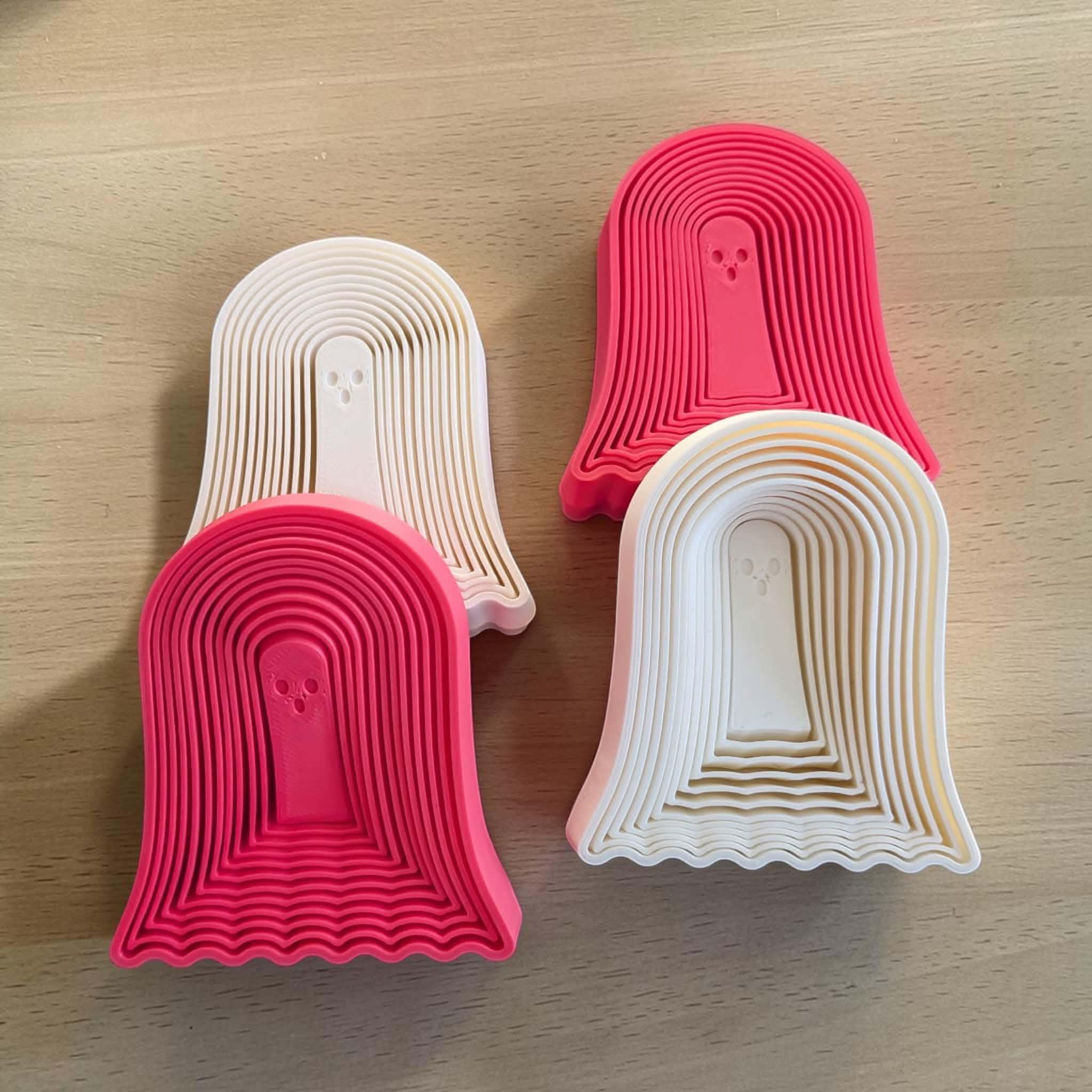 May include: Four ghost-shaped objects arranged in a group. Two are bright pink and two are off-white. Each object features a series of concentric arched ridges leading to a central ghost figure with two small circular eyes and a small mouth.